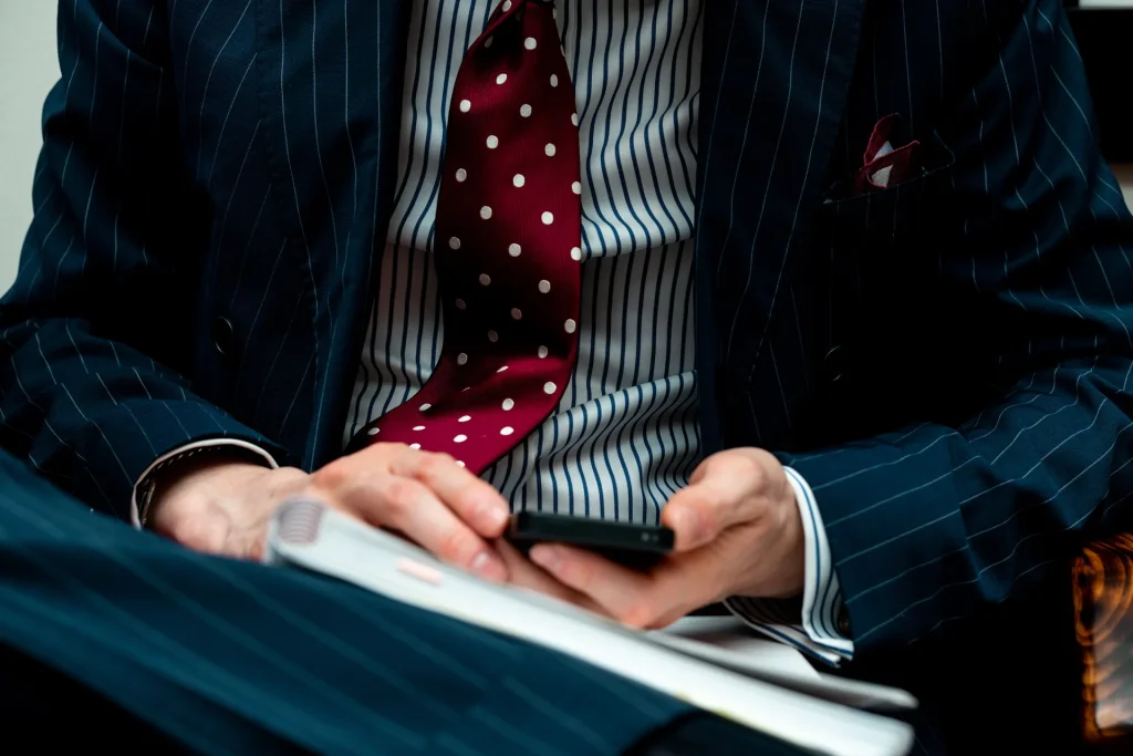 Manos de una persona con traje de raya diplomática y corbata granate que consulta el móvil sobre un cuaderno