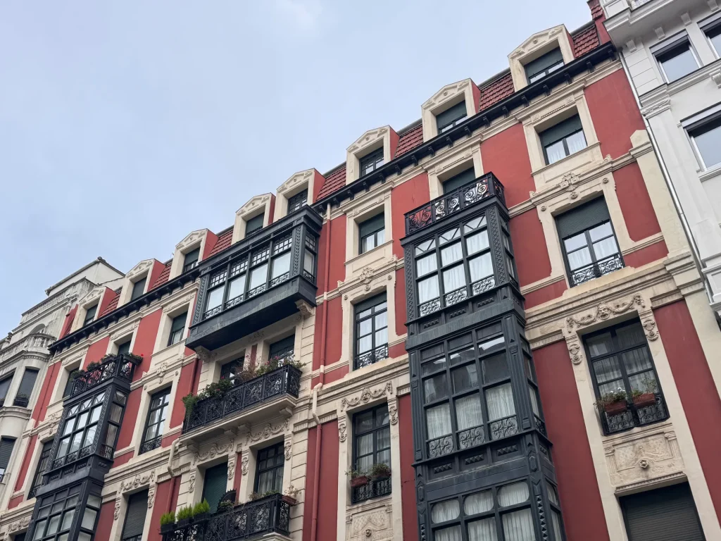 Fachada roja de un edificio histórico de Bilbao con miradores de hierro negro y balcones con flores